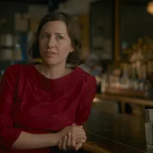 A woman in a red blouse sits leaning against a bar countertop, speaking to an interviewer behind the camera.