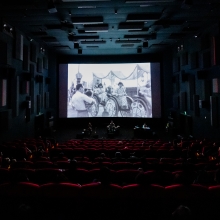 A large, dark theater screening “This Is Not a Film by Deng Nan-guang” with an experimental performance using drums and other instruments on the stage below the screen. 