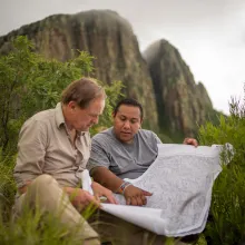 Two men look at a map in the wilderness against the background of a cliffside.