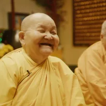 Two Buddhist nuns in a temple, with shaved heads, and goldish robes. The Buddhist Nun on the left is smiling big.