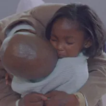 A bald man bends down to hug his daughter. The child is wearing a sad expression.