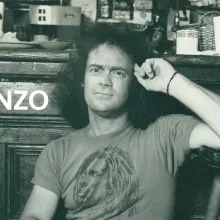 Black and white photograph of a man with dark, shoulder length hair leaning against a wooden cabinet