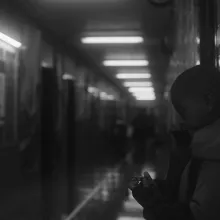 Black and white image of a child, photographed from the side, holding a watch while standing against the wall of a hallway 