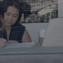 A woman with long hair and a dark top seen through a window writing in front of a laptop