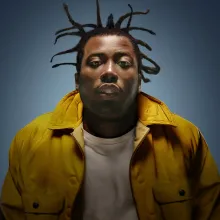 Photograph of a Black man with a yellow jacket, leaning into the camera.