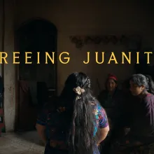 A woman from the back, sitting in front of other women, young and old, in a dark room. The woman in the center of the image has her hair braided any tied back with a white flower broche. She's wearing a colorful embroidered blue Maya huipil.