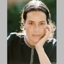 A woman with brown hair and wearing a black, colored shift leans her chin on one hand.