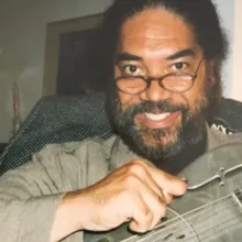 A man with dark hair and a beard, wearing glasses, smiles at the camera while holding a string instrument