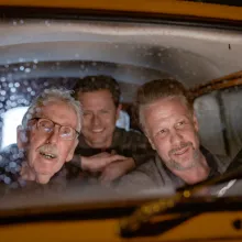 Three white men sit in a car looking out from the windshield 