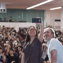 A young Asian woman and a middle-aged white man take a selfie in front of a large auditorium of spectators.