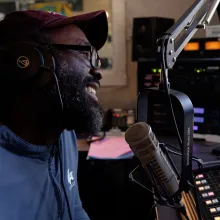 A black man sits in front of a microphone, smiling.