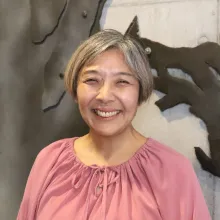 Headshot of Asako Fujioka. An east-Asian woman smiles at the camera with a concrete wall with black decorations behind her. She has straight graying hair and is wearing a loose dark pink blouse.