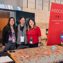 Three Asian American women stand behind a table of buttons, next to a red banner that reads "A-Doc."