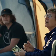 During golden hour, an older bespectacled astronomer looks up anxiously to the sky. Her younger colleagues, at a distance in a tent behind her, look to her. All are concerned.  