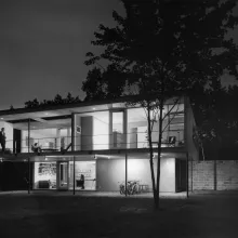 A black and white photo from the 1960's of a mid-century modern house at night.  It's an "upside down" house where the second story is larger than the first story so the house looks like two elegant rectangles stacked atop each other. The house is lit from the inside and a tall, daper man in a tuxedo stands on the upstairs terrace. A woman sits inside on the living room couch, and down below two young children can be seen.