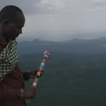 Black man holds a beaded staff and looks over a lush landscape