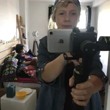 A white woman films herself with an iPhone. Behind her, on a bed, is her teenaged son.