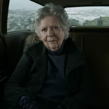 In a car interior, a white woman with short-cropped silver hair looks archly at the camera.