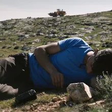 man reclines on a rock hill with a tank in the background