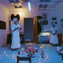 blue light colored a room of three balck women dressed in all white conducting a ritual while a group of people watched 