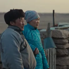 Two people dressed in traditional Mongolian clothes look into the distance.