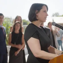 white woman standing at podium in black dress addressing crowd with people standin behind her