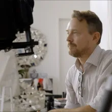 A white man sits behind some camera equipment in a dressing room.