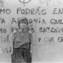 Black and white photo of a woman leaning against a wall. She is a wearing a striped tank top, and her hands are in her pockets. 