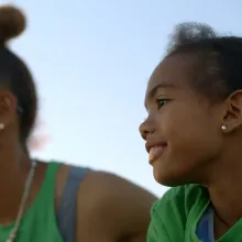 Wearing similarly-colored green shirts, a Black woman and her daughter smile while looking off-screen.