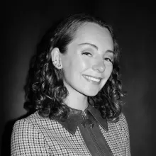 Headshot of Berry Hahn. A black and white image of a smiling mixed-race woman with mid-length curly brown hair, wearing a jacket with a houndstooth pattern.