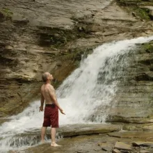 A shirtless man stands in front of a waterfall with his arms spread