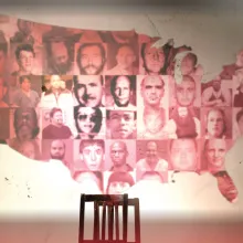 Map of the lower 48 states of the US projected on a wall with a chair in front of it.  Each state of the map has a mugshot or face of a mass shooter.