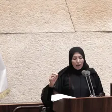 A woman in a black hijab delivers a speech at the podium of the Israeli Knesset, standing next to a large Israeli flag.