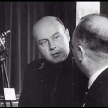 A bald white man, in a dark uniform, talks to another man with his back facing the camera.