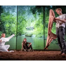 In front of an illuminated screen of the Amazon, three actors sit or stand in the dirt.