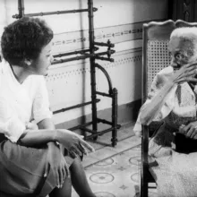 Two Black women sit on whicker chairs, talking cordially to each other.