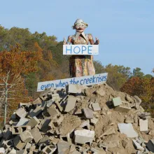 Atop a pile of concrete rubble, there is a statue of a person holding a sign that says "Hope... Even when the creek rises..."