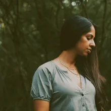 A dark-skinned woman with long black hair wearing a sage green dress looks downward as she is surrounded by trees.