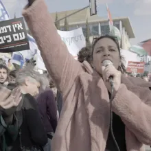 In a crowd of protestors, a woman in a pink coat speaks into a microphone while raising her right arm.