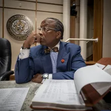 A Black man in a navy suit sits at a wooden courthouse table.