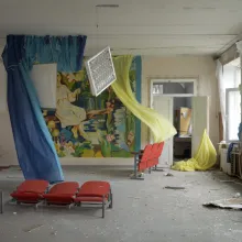 An empty room that has been bombed with fabrics, chairs strewn around and windows open.