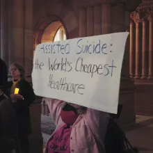 A woman in a pink jacket holds up a sign that reads: "Assisted Suicide: The World's Cheapest Healthcare."