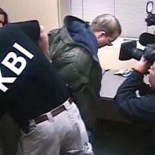 Lo-res still image of a man in a green jacket being handcuffed by a man wearing a "KBI" shirt. There is another man with a large digital video camera filming the face of the man being arrested, and a sliver of a boom pole in another corner of the image.