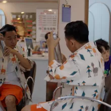 A young Chinese man in a colorful button-up shirt sits in front of a mirror in a barbershop, spraying hairspray onto his head.