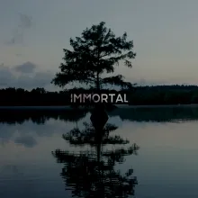 A swamp at dusk surrounded by trees and in the center of the swamp is a tree standing in the middle of the water. The tree's reflection in the water is directly beneath the tree in the middle of the frame. Centered in the image is the word IMMORTAL.