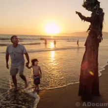 A man on stilts dressed as a tree waves to little boy and his father on the beach at sunset