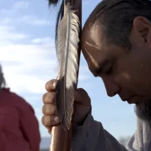 Close up of a man praying, with a woman standing in the background.