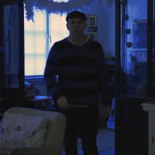 A man in a striped sweater and cap stands in a dimly lit room bathed in a cold blue tint, captured in the early morning light of winter. 