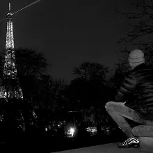 A figure wearing a dark puffy jacket looks at the lit-up Eiffel Tower in the background.