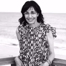 In black and white, Andrea Nevins stands on a balcony with the ocean behind her, smiling brightly.. 
